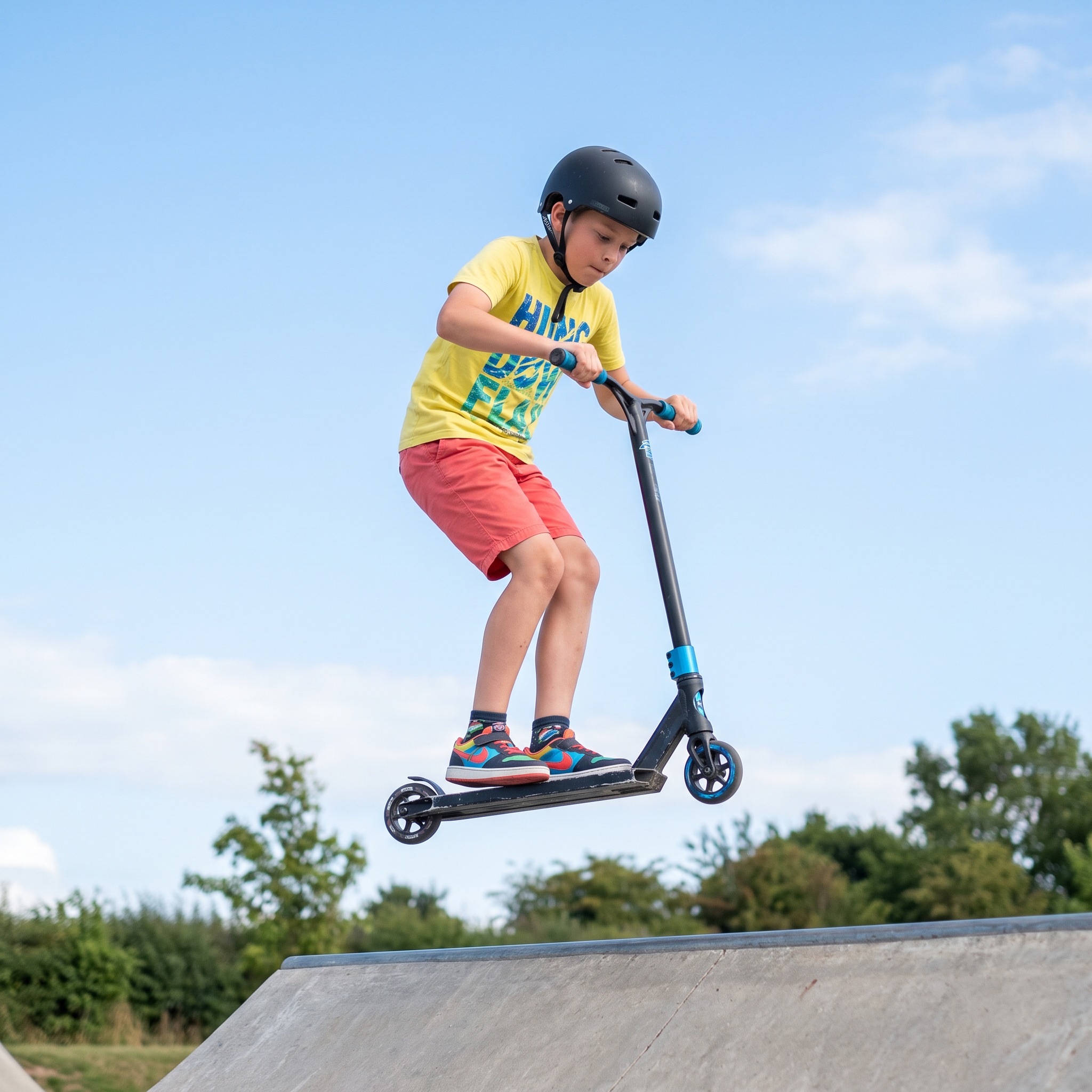 A child mid-air on a stunt scooter above a skate park ramp, wearing a helmet
