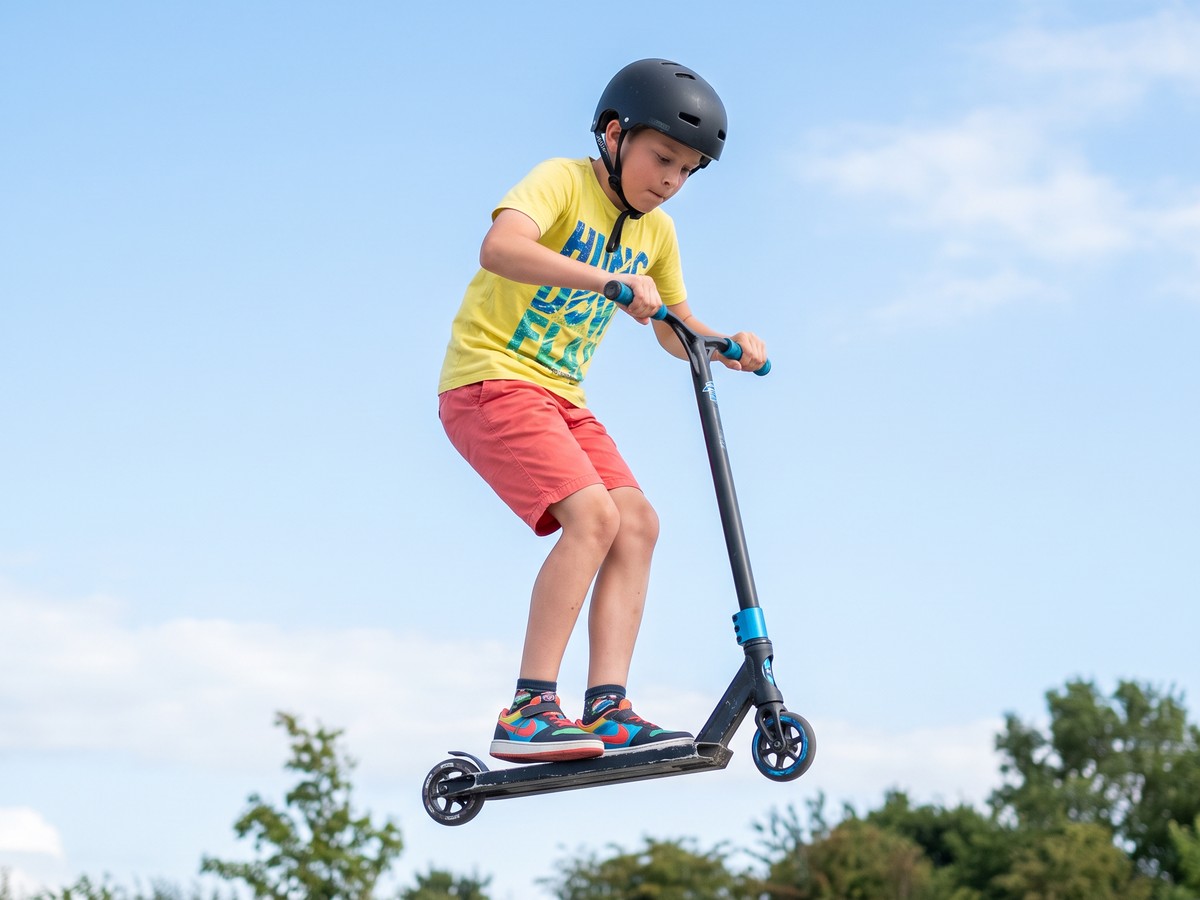 A child mid-air on a stunt scooter above a skate park ramp, wearing a helmet